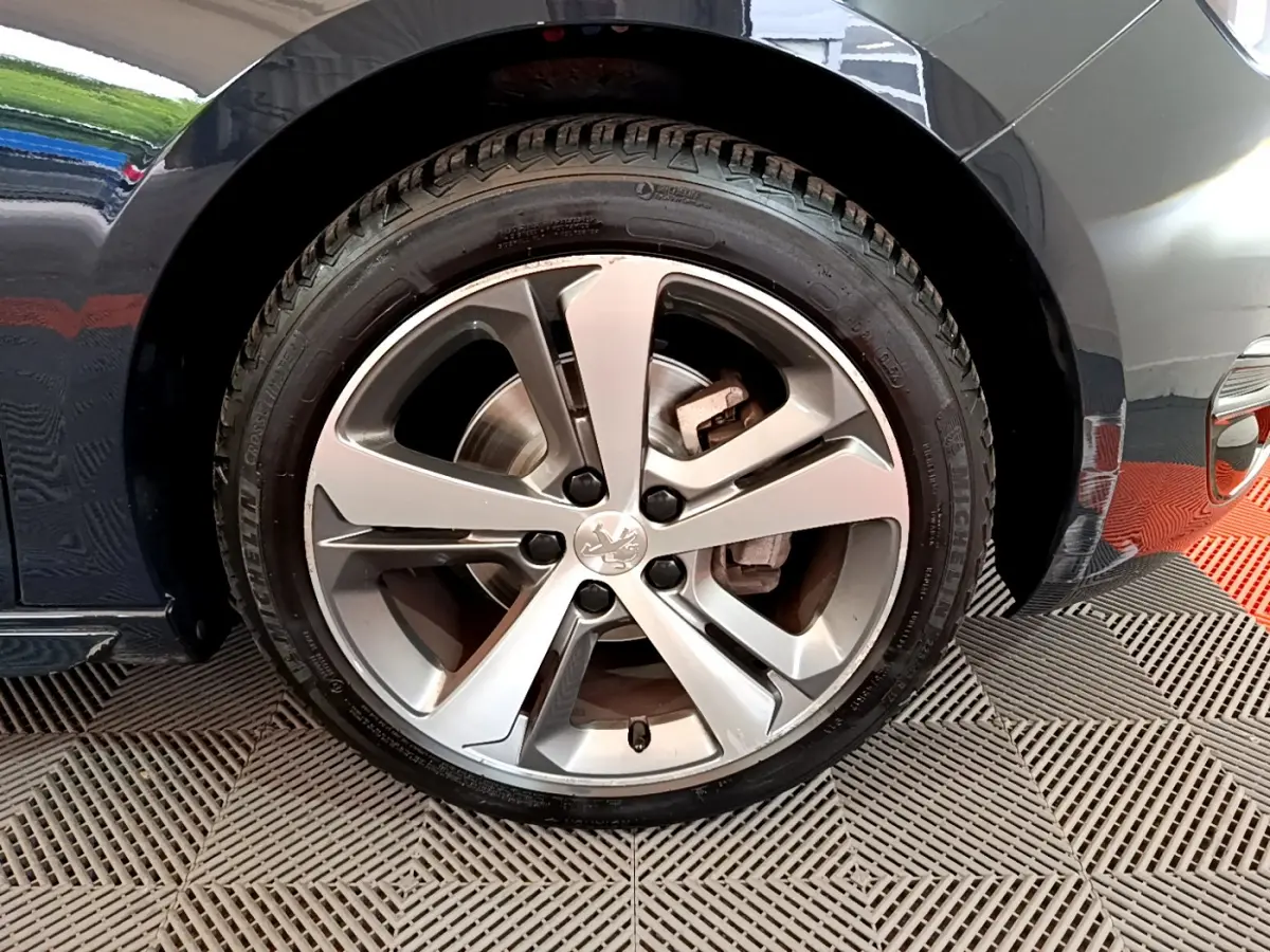 Gros plan sur la roue avant droite d'une Peugeot 308 gris foncé, mettant en valeur la jante alliage et le logo lion central.