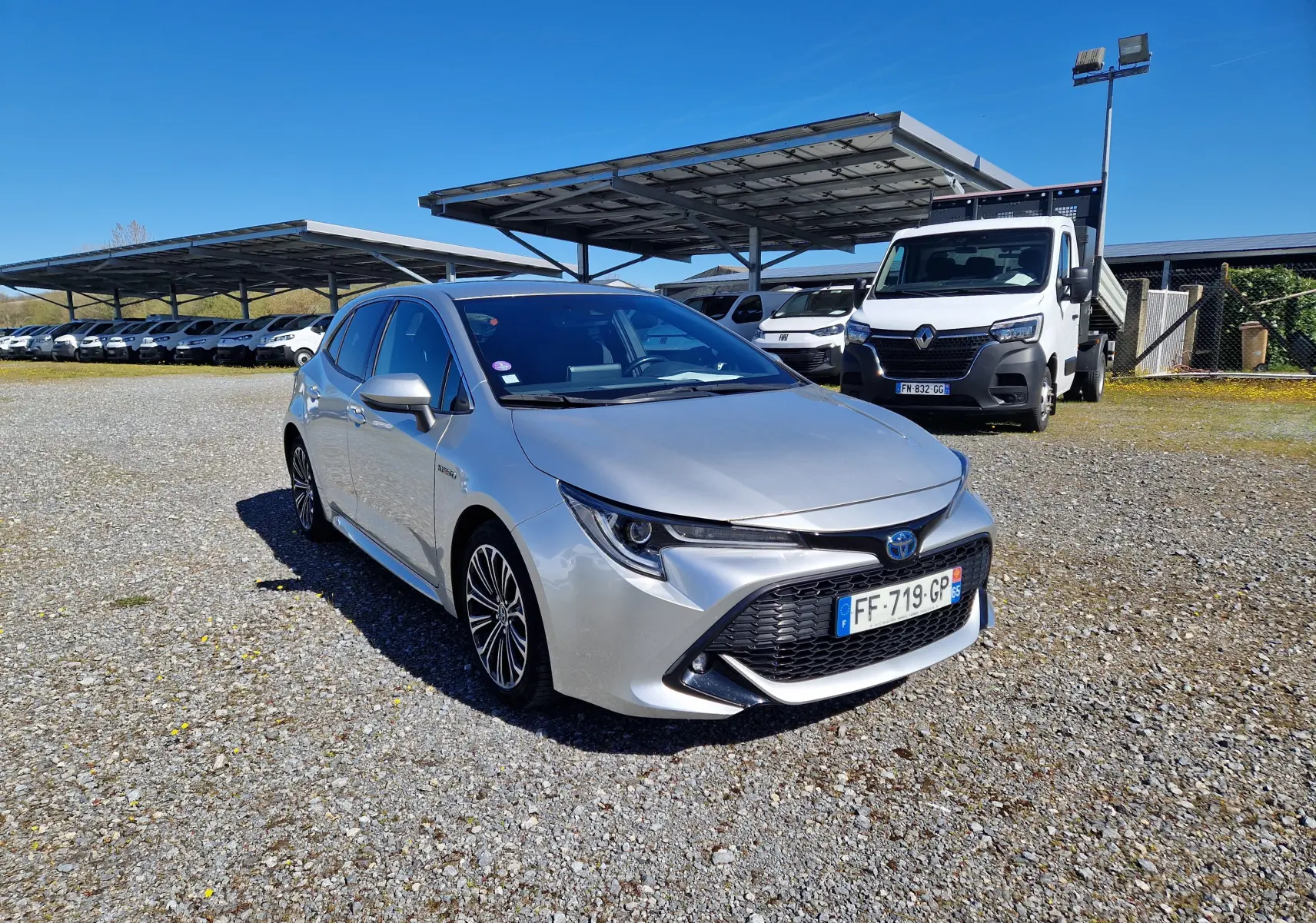 Toyota Corolla hybride gris clair métal vue 3/4 avant droit sur parking extérieur avec plusieurs véhicules en arrière-plan.