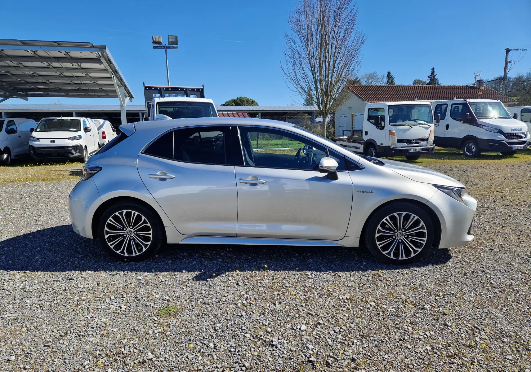 Vue de profil côté gauche d'une Toyota Corolla hybride gris clair métal avec jantes alliage et badge hybride visible.