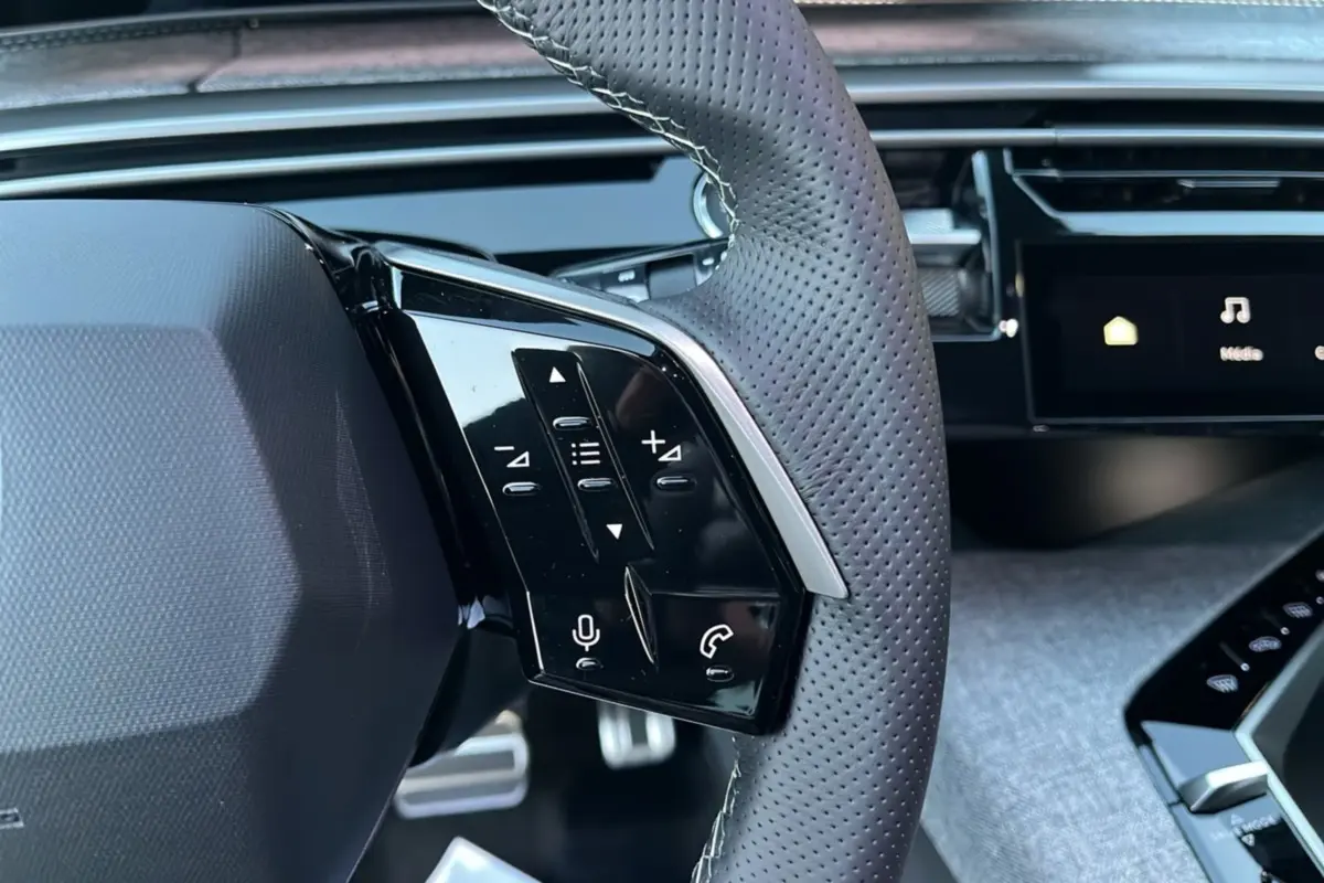 Gros plan sur le volant cuir perforé gris du Peugeot 5008 Hybrid 145 GT avec commandes multifonctions et pédalier aluminium visible.