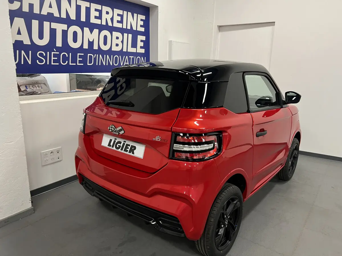 Vue 3/4 arrière droite de la LIGIER JS50 BEV 8KWH ELITE rouge avec toit noir et jantes alu noires en intérieur showroom.