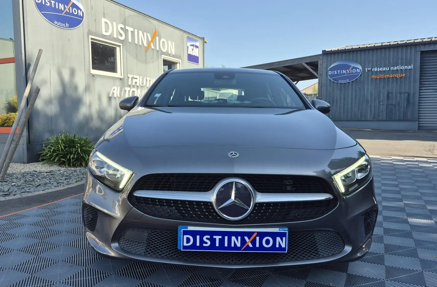 Vue frontale d'une Mercedes Classe A 180d gris foncé avec calandre noire et phares LED allumés, devant un bâtiment Distinxion.