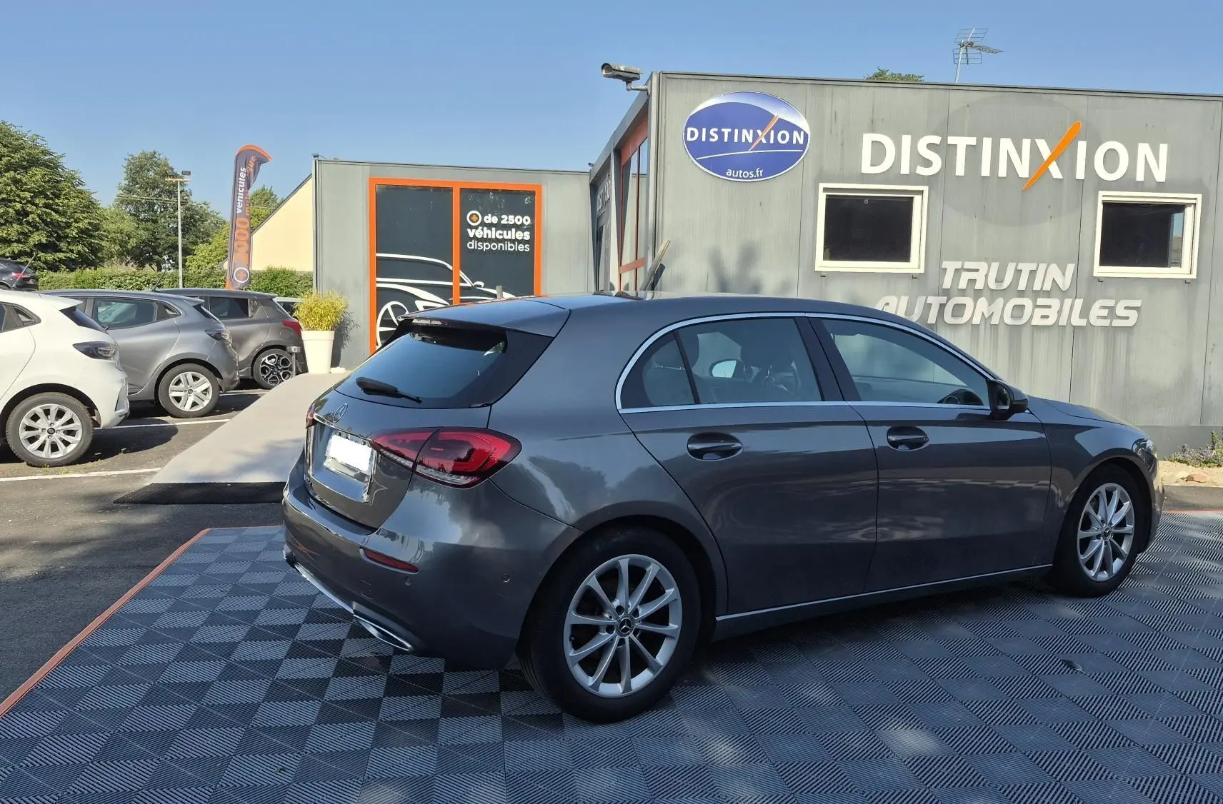Mercedes Classe A 180d gris foncé vue 3/4 arrière droit, stationnée devant un bâtiment avec logo Distinxion.