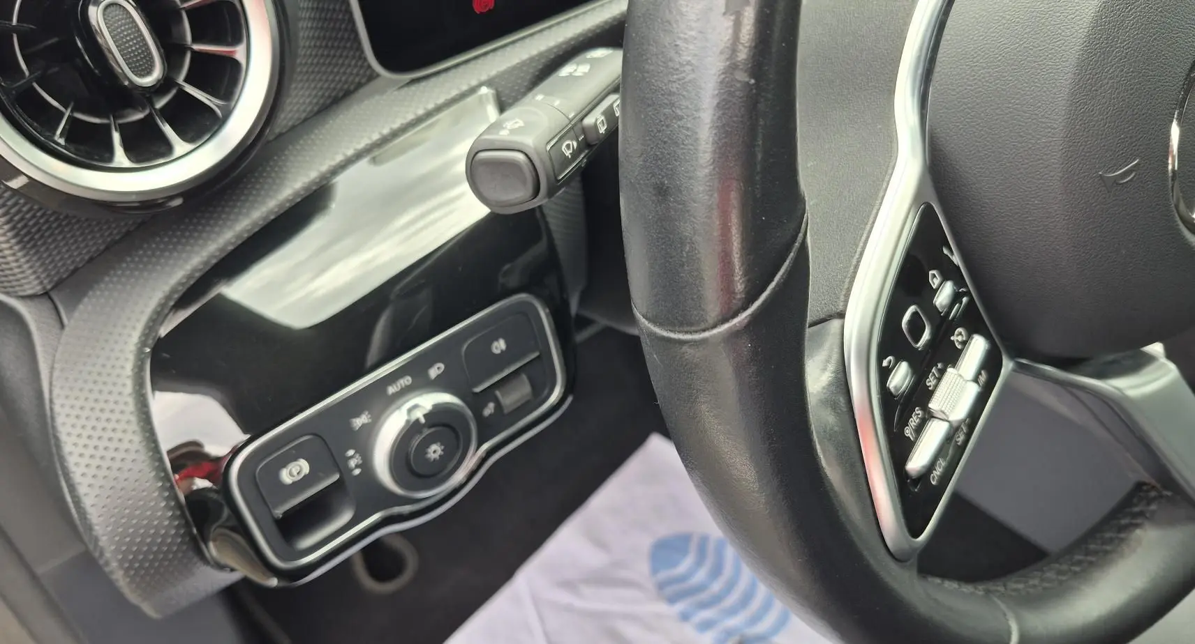 Vue rapprochée de l’intérieur côté gauche du volant cuir noir d’une Mercedes Classe A 180d gris foncé, avec commandes éclairage.