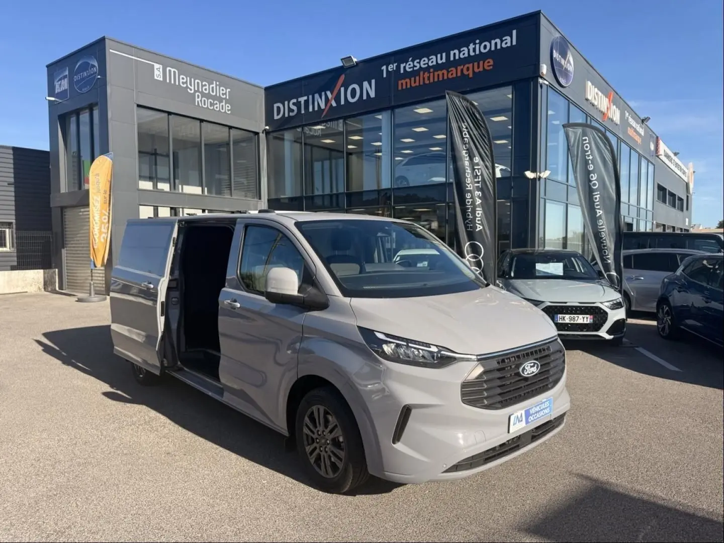 Ford Transit Custom Fourgon gris en 3/4 avant droit, porte latérale coulissante ouverte, devant un bâtiment commercial.
