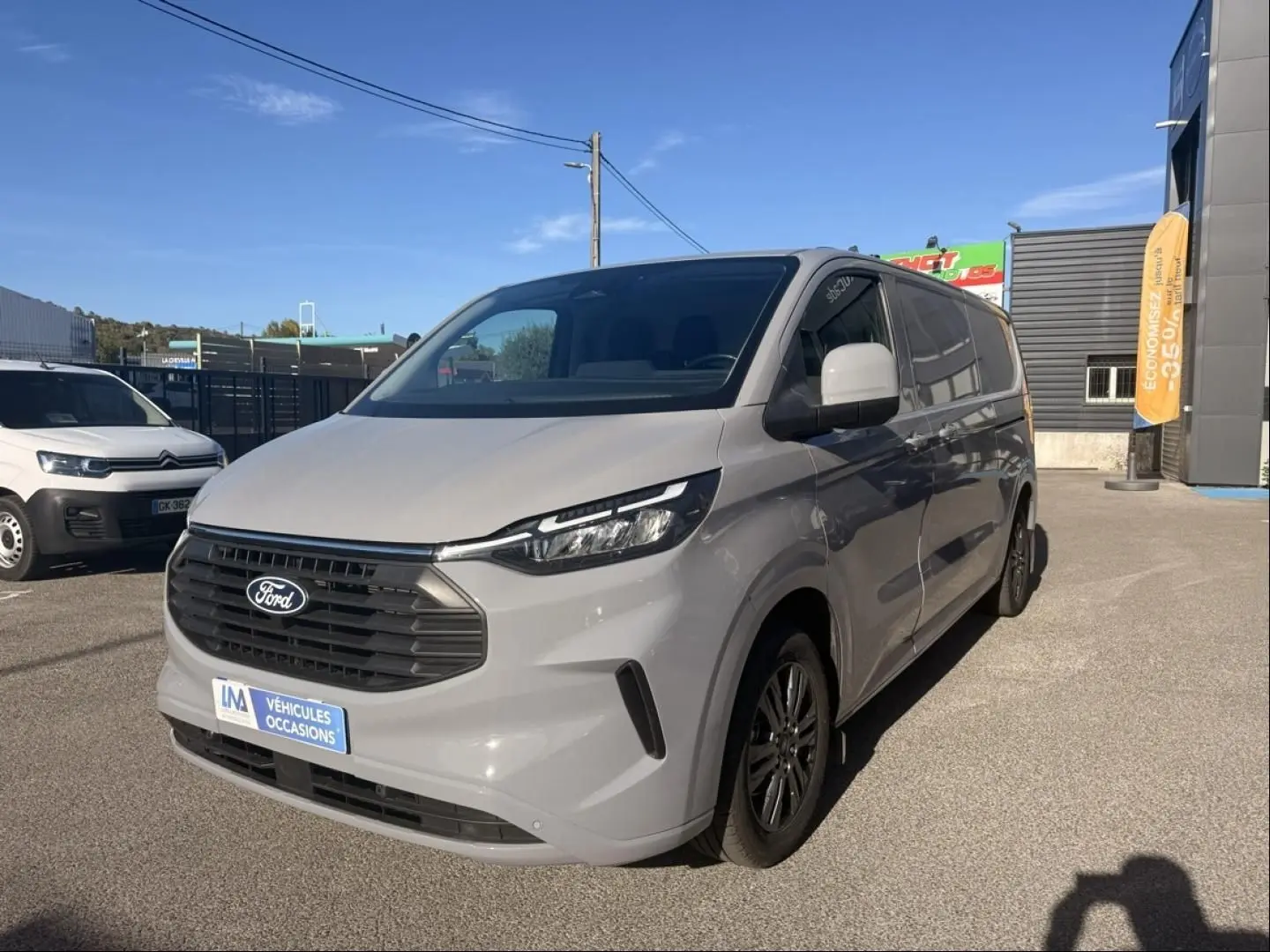 Ford Transit Custom Fourgon gris en 3/4 avant droit, avec calandre noire et jantes alliage sombres, stationné en extérieur.