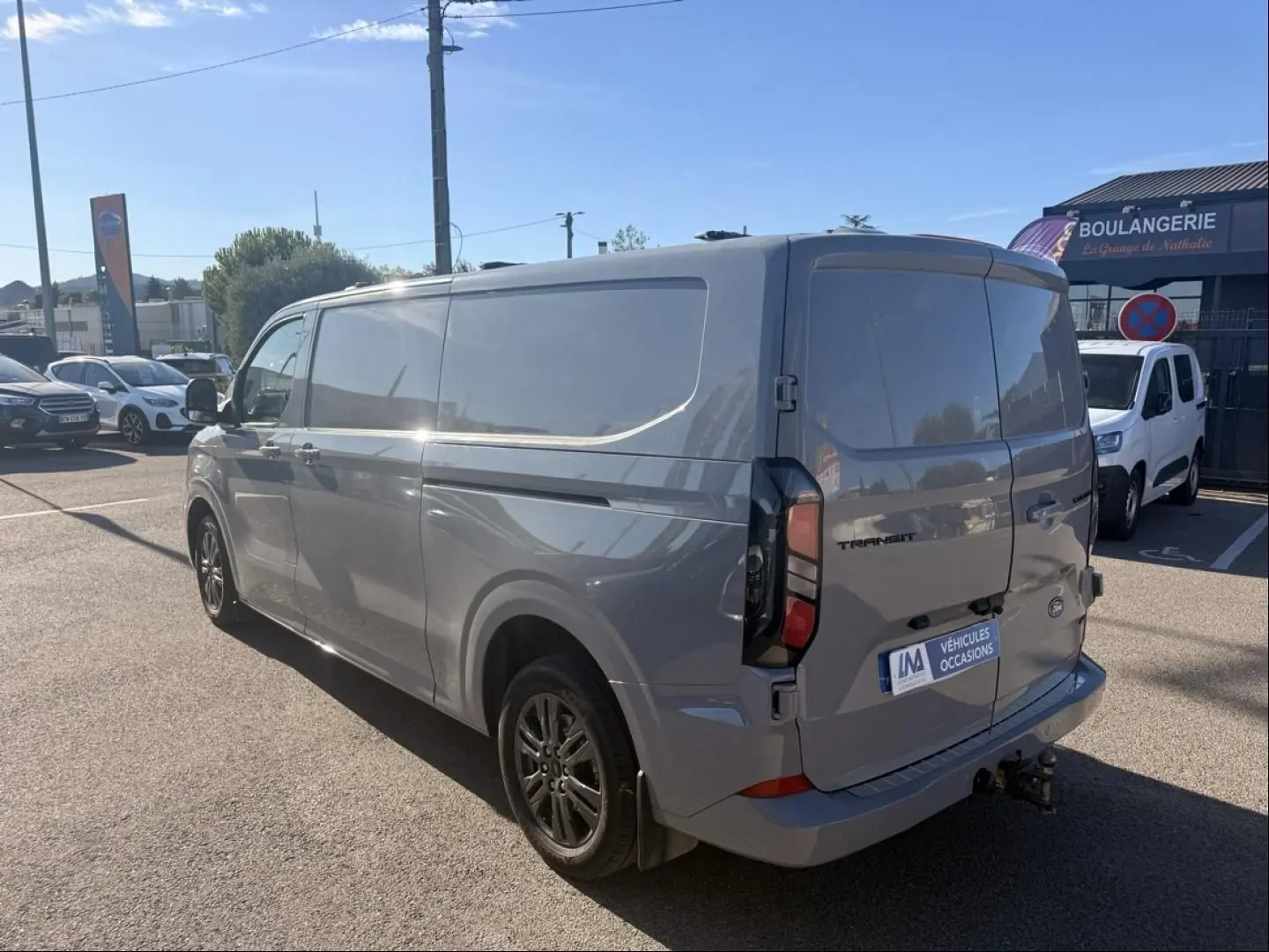 Vue 3/4 arrière droite d’un Ford Transit Custom Fourgon gris 2024 avec hayon et attelage visible.
