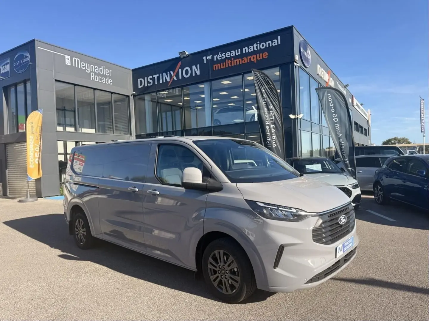 Ford Transit Custom Fourgon gris en 3/4 avant droit devant un showroom multimarque par temps clair.