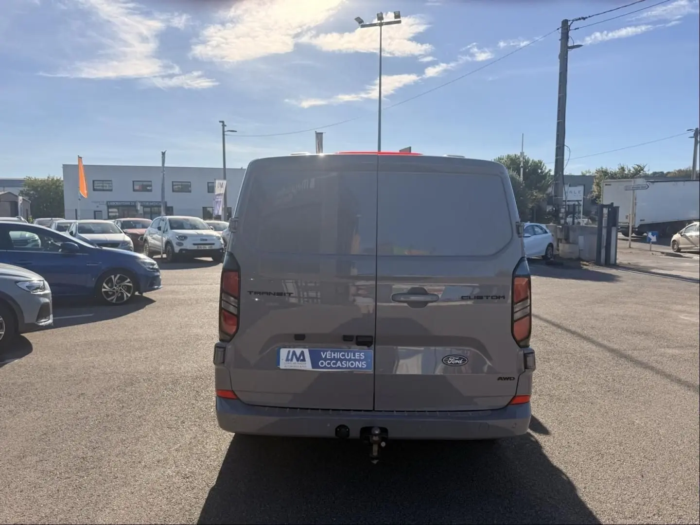 Vue arrière d’un Ford Transit Custom gris 2024 avec hayon fermé et attelage visible sur parking extérieur.