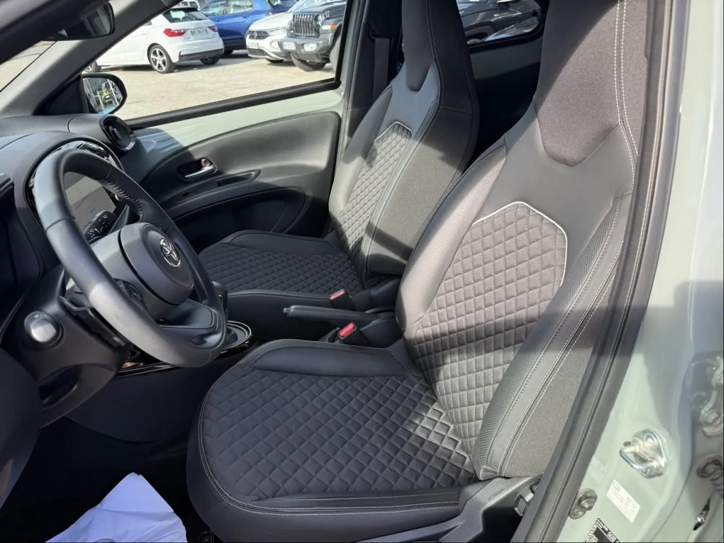 Vue intérieure côté conducteur de la Toyota Aygo X 2025, sièges noirs avec surpiqûres losangées et volant multifonction.