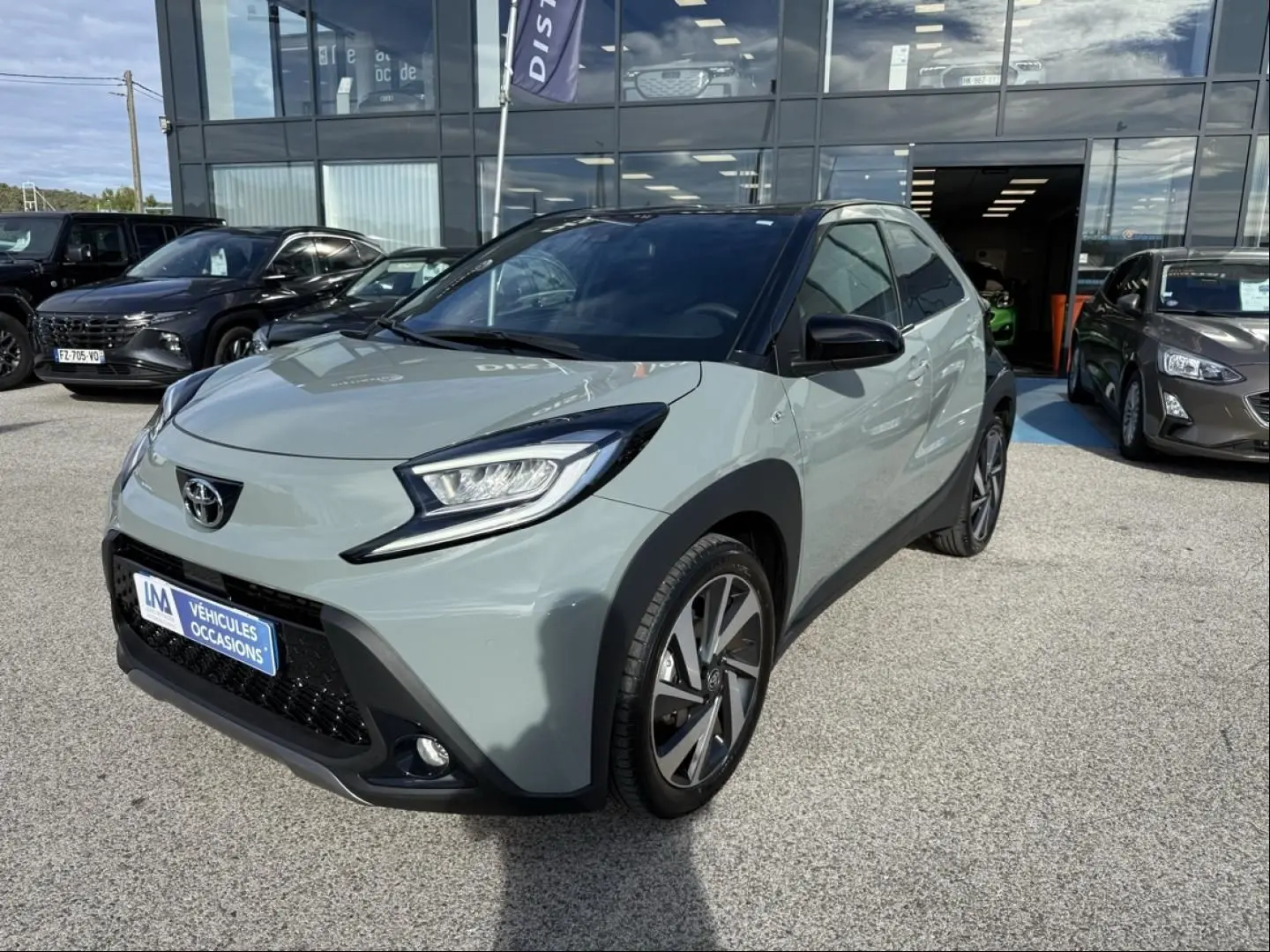 Toyota Aygo X 2025 vue 3/4 avant en vert aventurine avec toit noir, garée devant un showroom.
