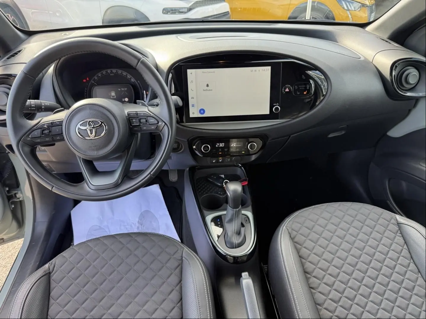 Vue intérieure frontale du tableau de bord et volant de la Toyota Aygo X 2025, avec écran tactile et sellerie grise matelassée.