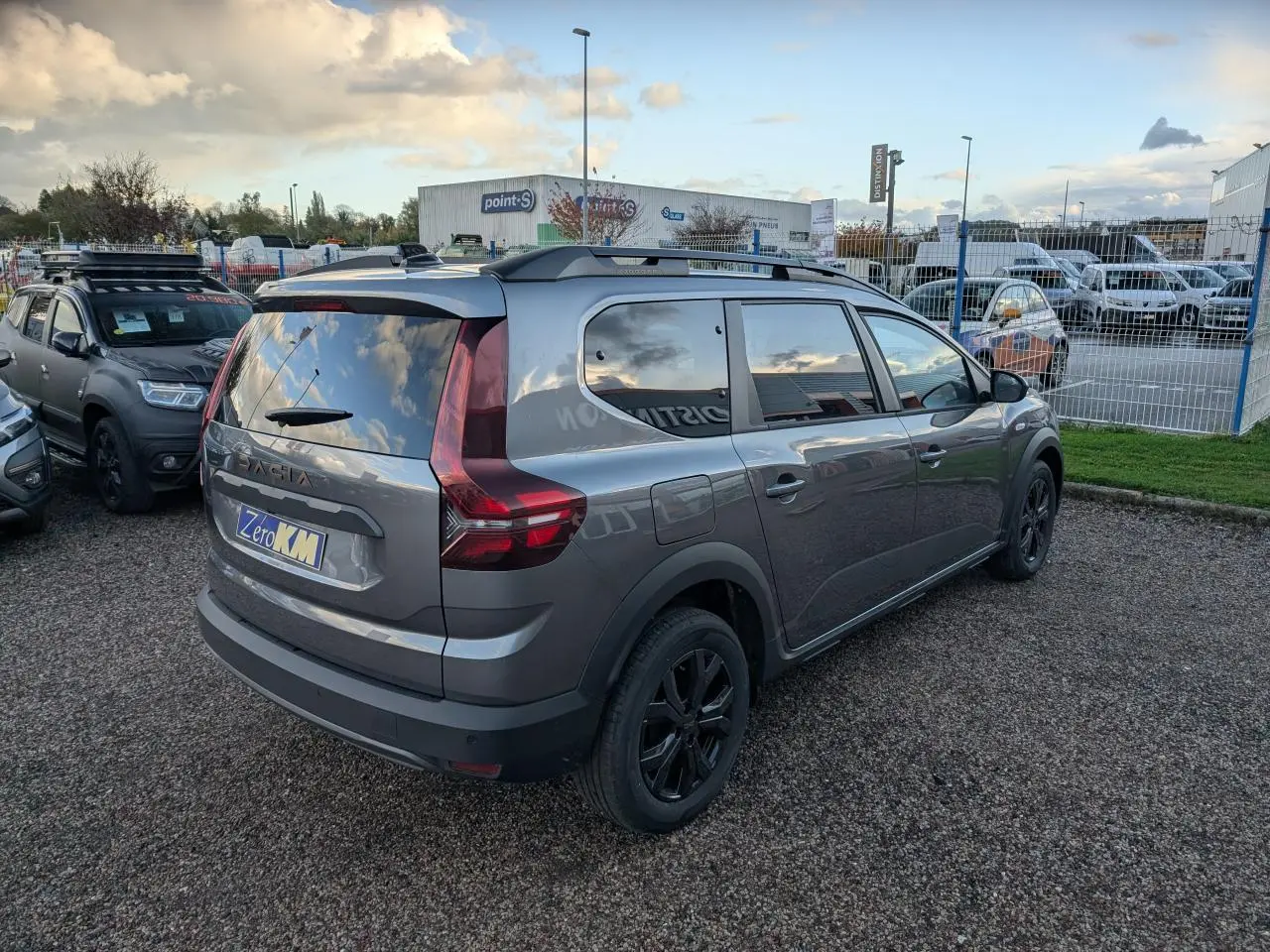 Vue 3/4 arrière droite d’un Dacia Jogger gris métallisé avec jantes noires et barres de toit, stationné sur un sol gravillonné.