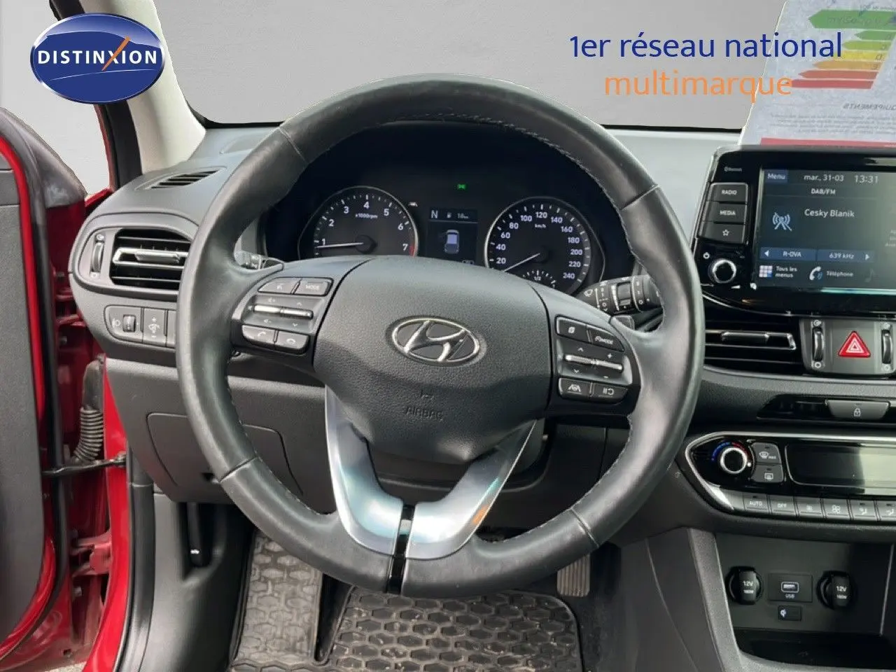 Vue intérieure centrée sur le volant cuir de la Hyundai i30 SW 2023 rouge Sunset Red Metal avec tableau de bord et écran tactile.