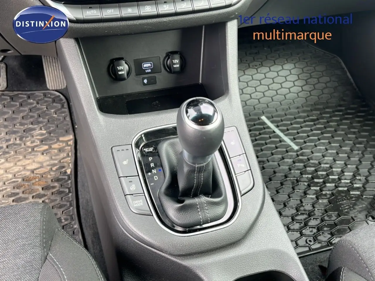 Vue rapprochée de la console centrale de la Hyundai i30 SW 2023, avec levier de boîte automatique et tapis caoutchouc noir.