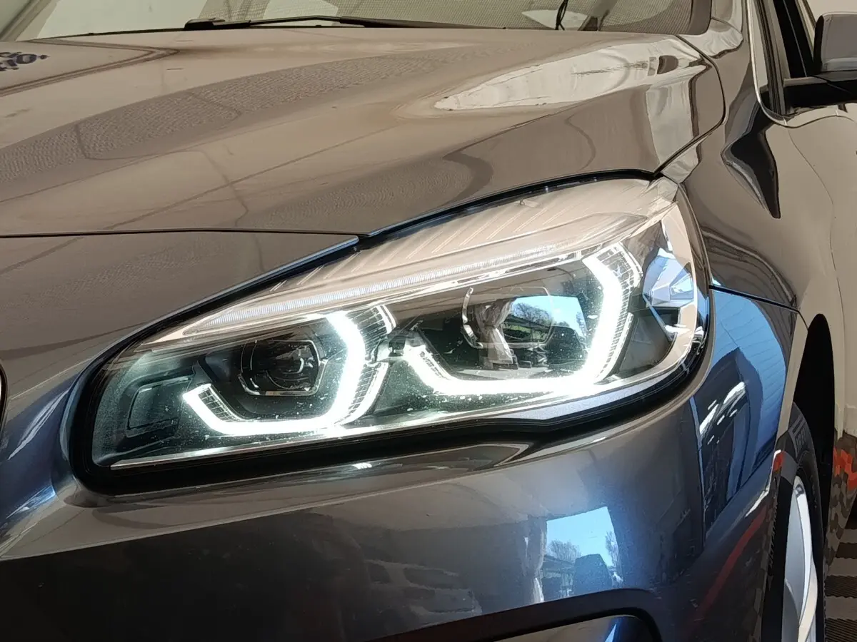 Gros plan sur le phare avant droit allumé d'une BMW Série 2 Gran Tourer F46 gris clair, reflet sur la carrosserie.