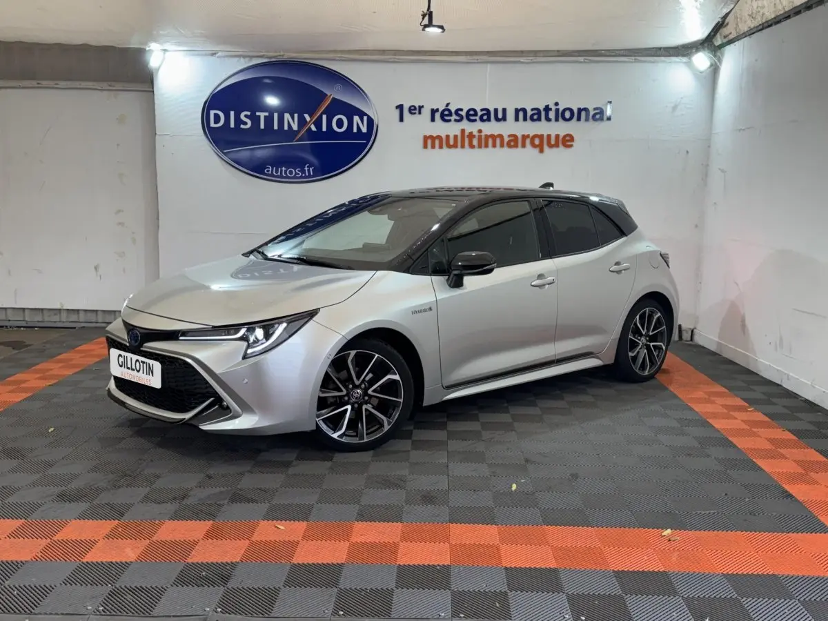 Toyota Corolla Hybride gris clair vue 3/4 avant droit, avec jantes alliage et phares LED allumés.