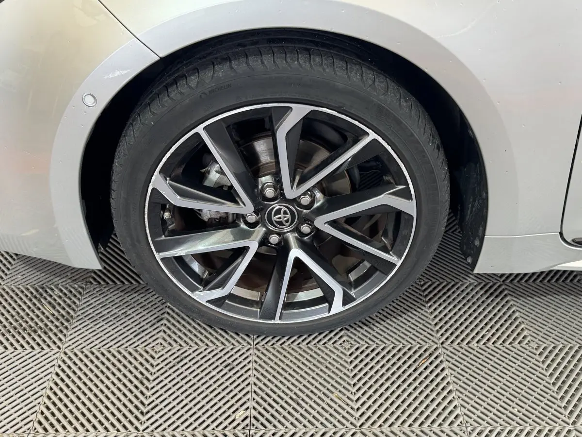 Gros plan sur la roue avant gauche de la Toyota Corolla hybride gris clair, avec jante alliage noire et argentée au design moderne.