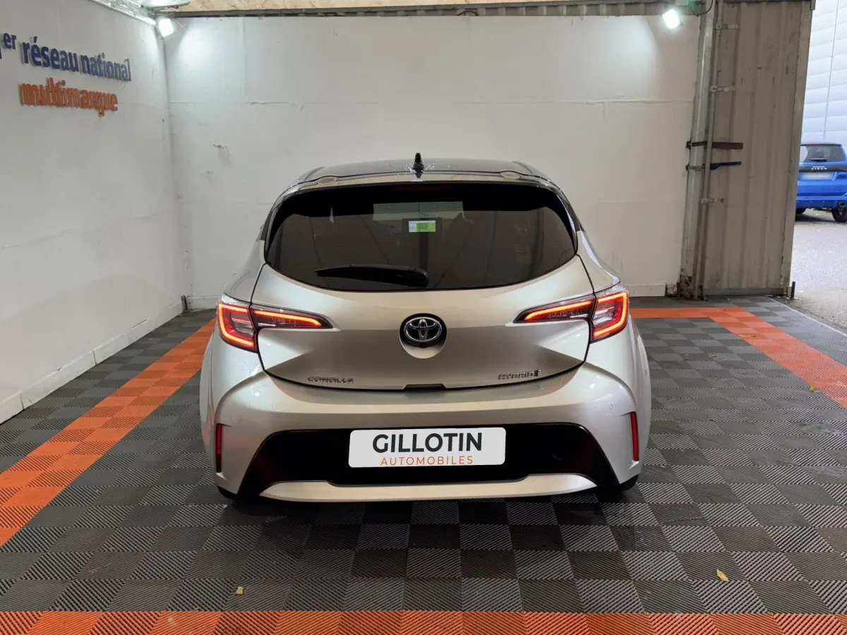 Vue arrière d'une Toyota Corolla Hybride gris clair, feux LED allumés, dans un garage avec sol à damier noir et orange.