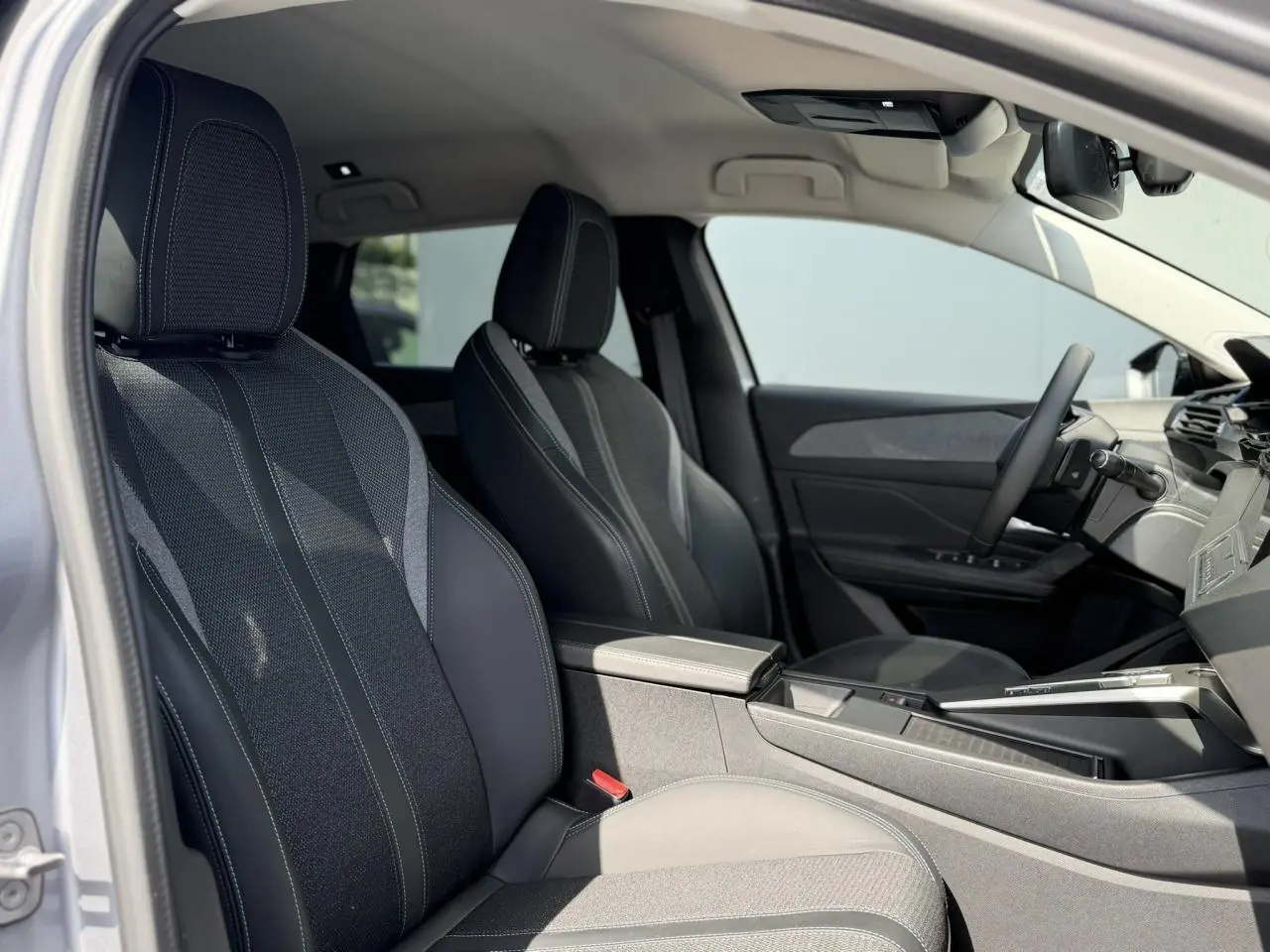 Vue intérieure côté conducteur de la Peugeot 308 SW 2025, sièges tri matières gris avec surpiqûres bleues et accoudoir central.