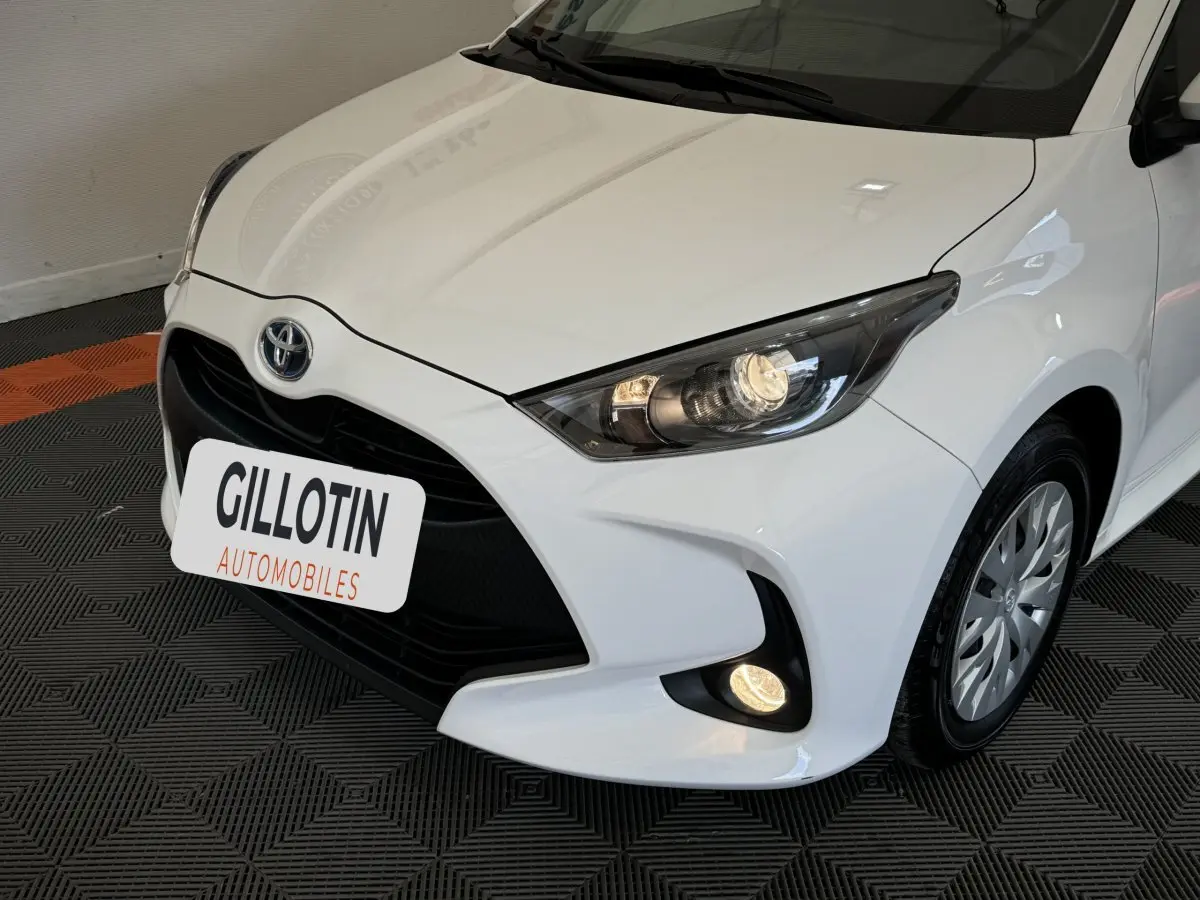 Vue 3/4 avant droite d'une Toyota Yaris hybride blanche avec phares allumés et calandre noire distinctive.