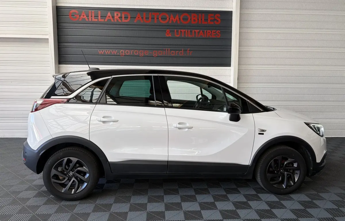 Vue de profil côté gauche d'un Opel Crossland blanc avec toit noir et jantes foncées, stationné en intérieur.