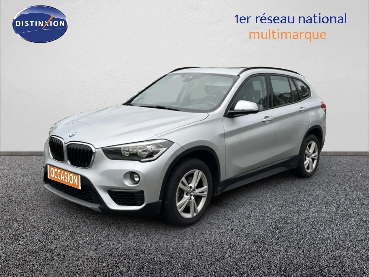 BMW X1 18i gris argent métal en 3/4 avant droit, avec jantes alliage et calandre noire distinctive.