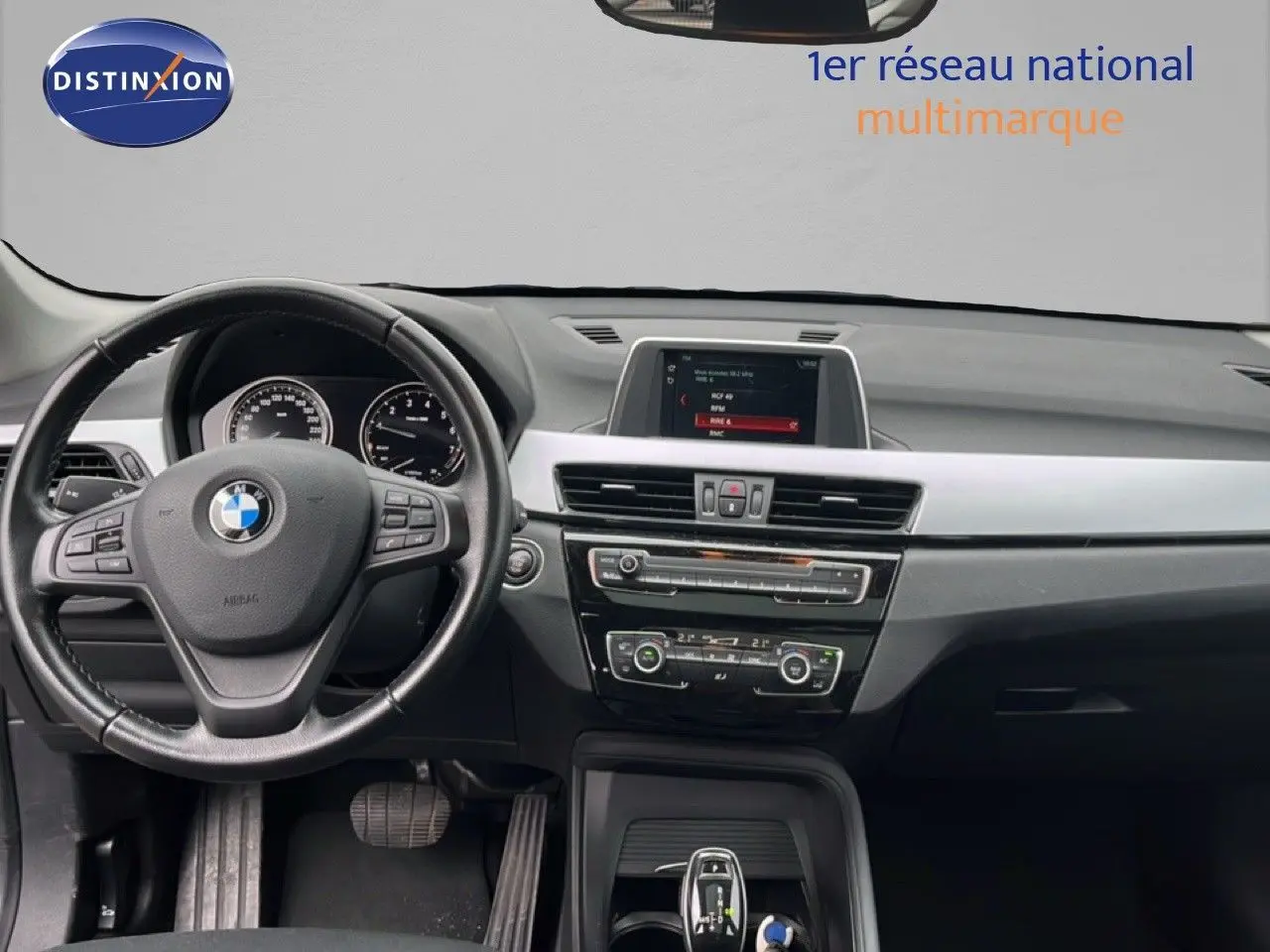 Vue intérieure centrée sur le tableau de bord et le volant noir d'une BMW X1 18i 2018, avec écran multimédia allumé.