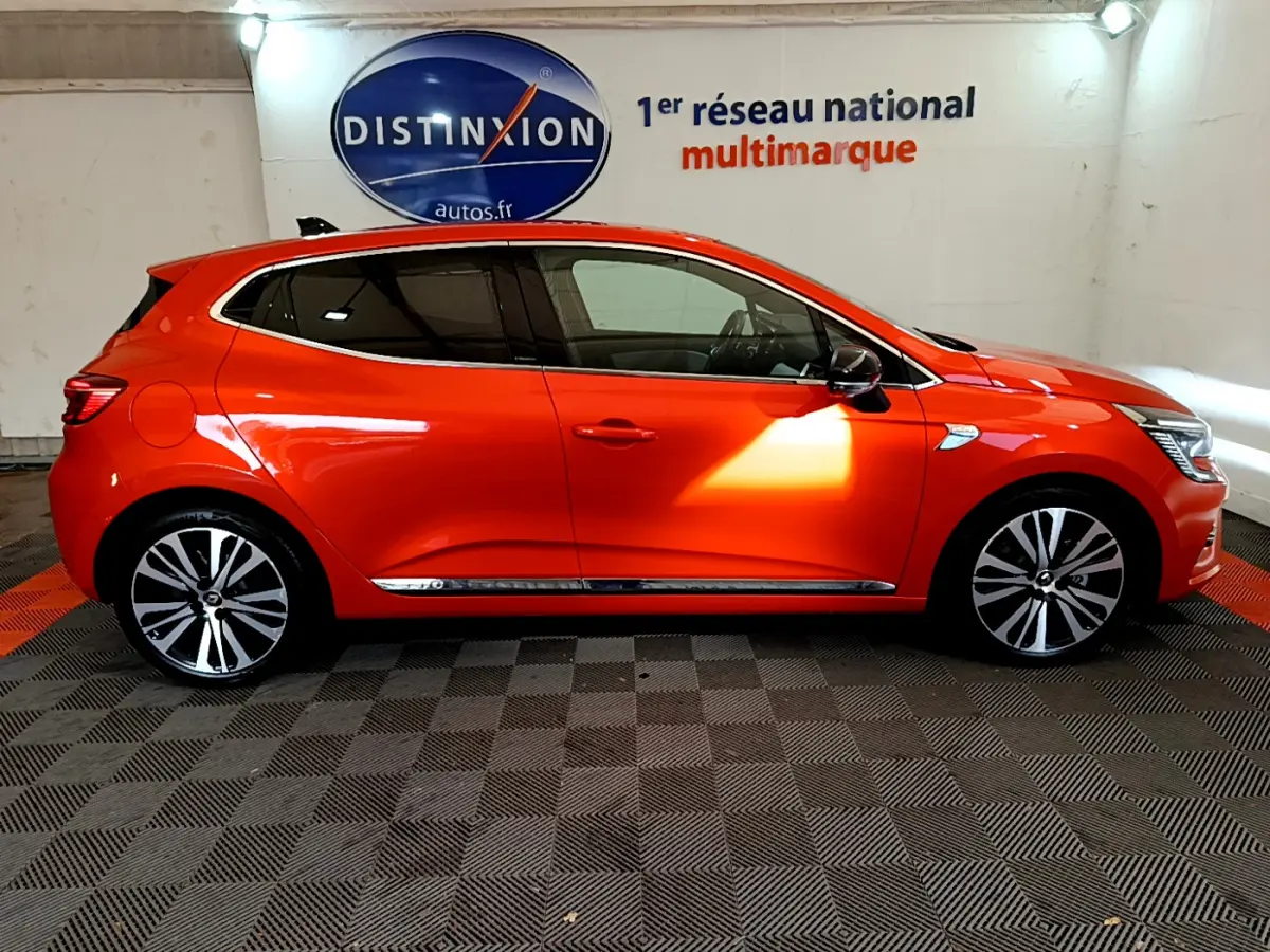 Profil droit d'une Renault Clio Initiale Paris E-TECH 140 orange, avec jantes alliage et vitres teintées, en intérieur showroom.