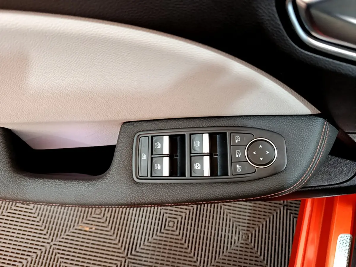 Gros plan sur la commande des vitres côté conducteur avec surpiqûres orange, intérieur clair et carrosserie orange visible.