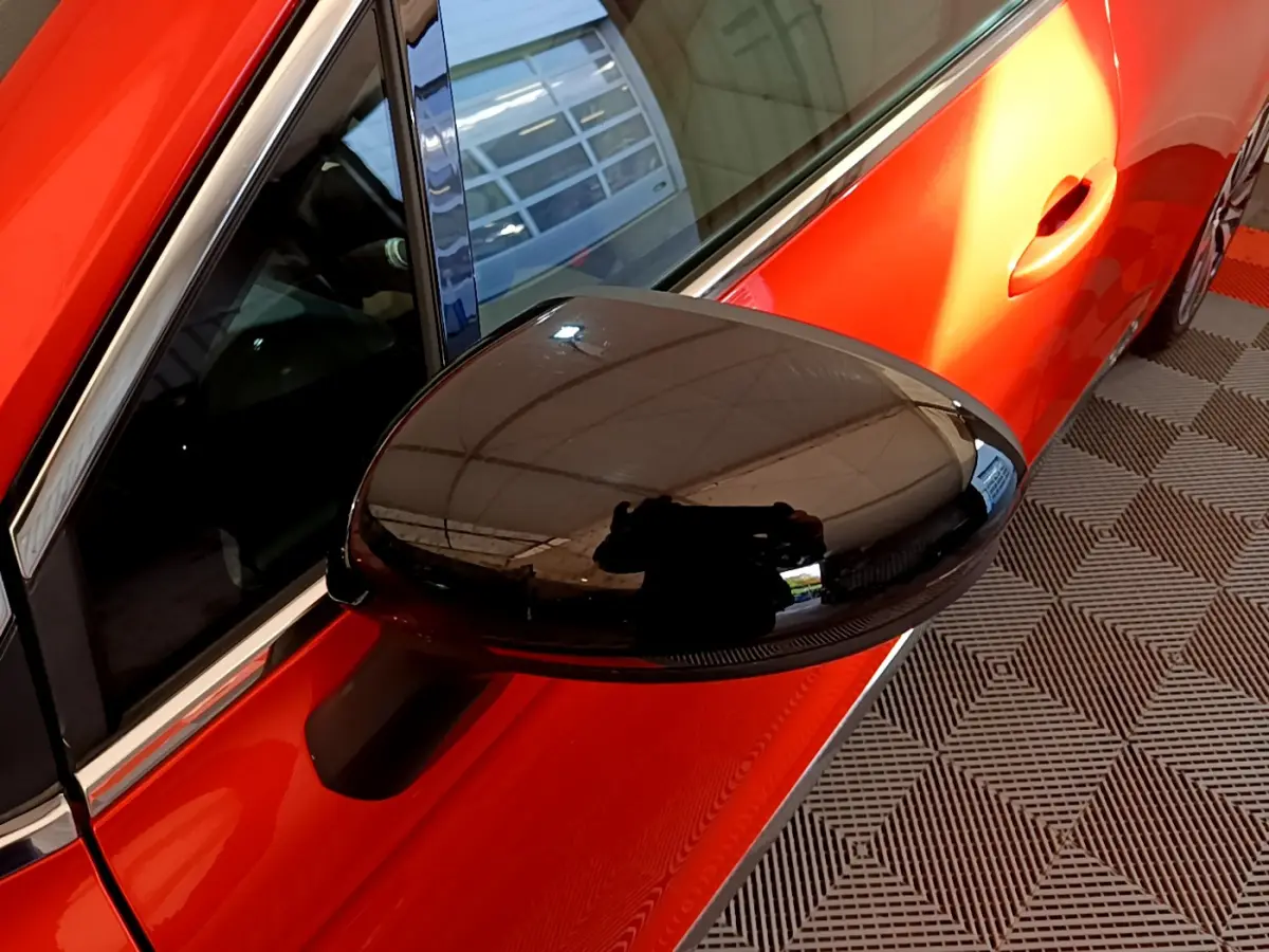 Rétroviseur noir brillant côté gauche d'une Renault Clio orange vue en gros plan sur fond de sol à motifs.