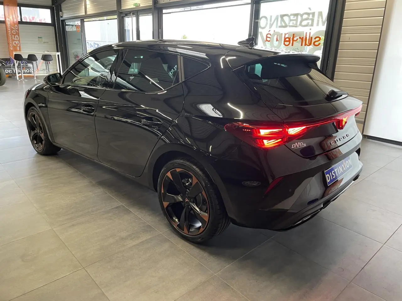 Vue 3/4 arrière droite d'une CUPRA Leon noire avec jantes alliage cuivrées et feux arrière allumés en intérieur showroom.