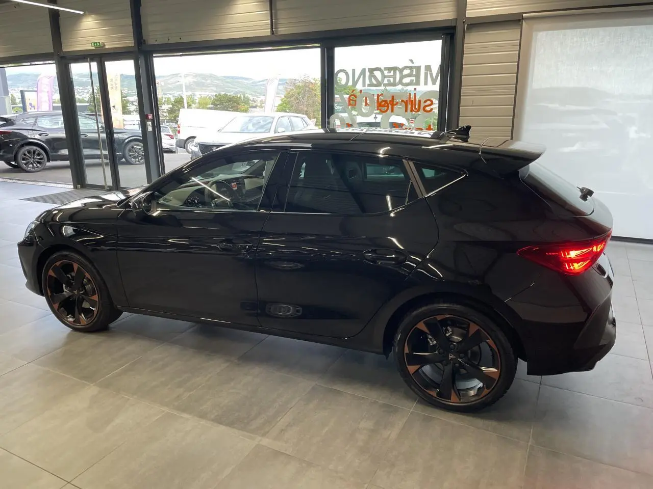 Vue de profil côté gauche d'une CUPRA Leon noire avec jantes alliage cuivrées dans un showroom lumineux.