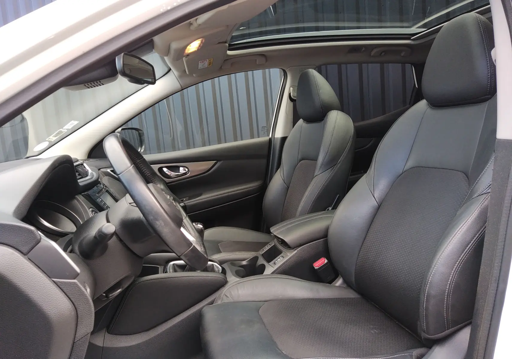 Vue intérieure côté conducteur du Nissan Qashqai blanc 2018, sièges cuir noir et toit ouvrant panoramique visible.