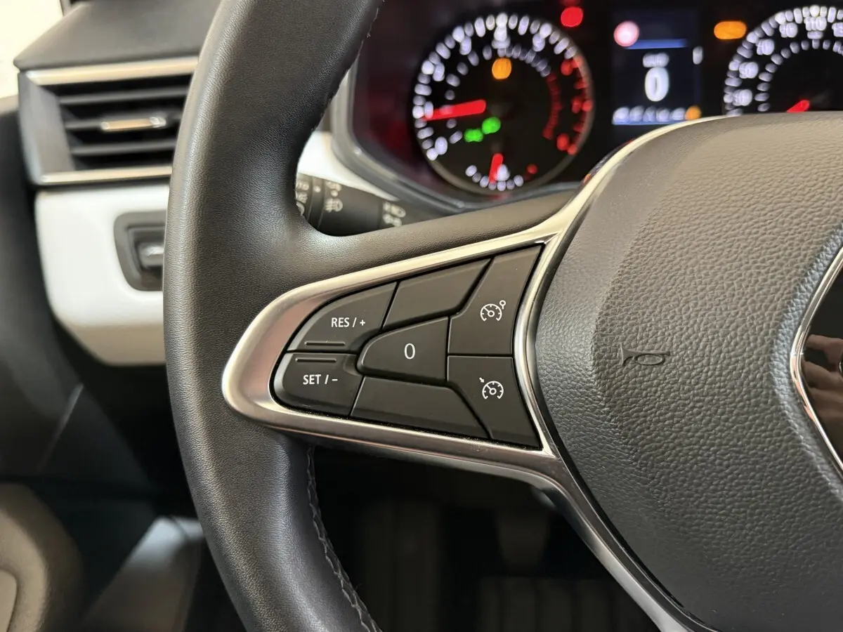 Gros plan sur le volant cuir synthétique de la Renault Clio blanc, avec commandes de régulateur de vitesse et tableau de bord flou.