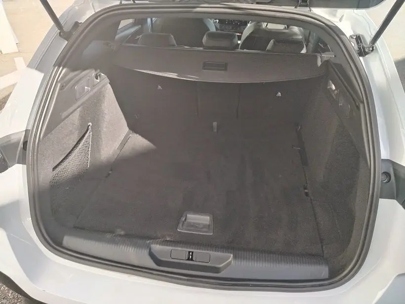 Coffre ouvert vu de l'arrière d'une Peugeot 308 SW Hybrid 145 blanc Okenite, espace de chargement spacieux et tapis noir.