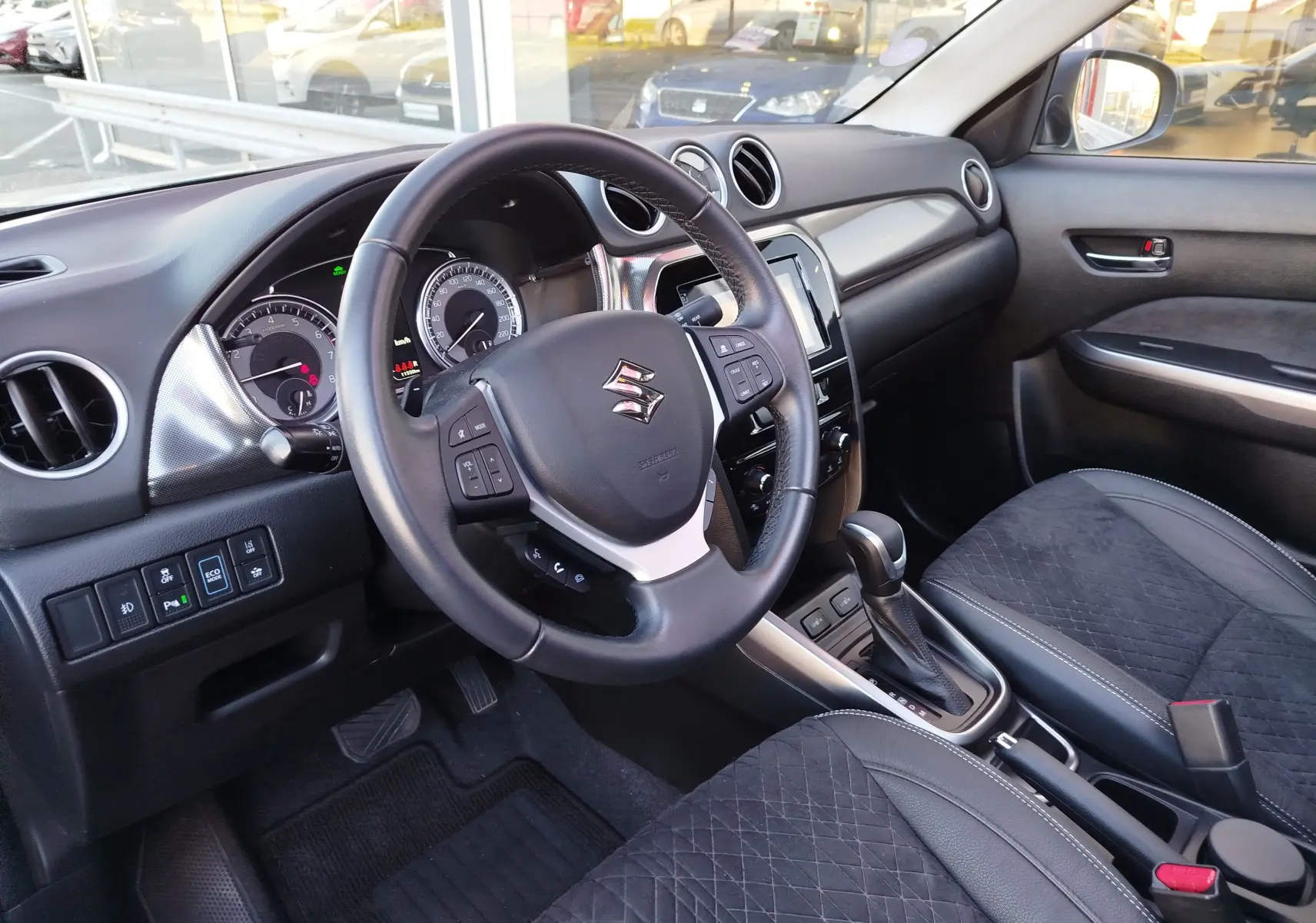 Vue intérieure côté conducteur du Suzuki Vitara 2023, volant cuir, tableau de bord noir et sièges tissu noir avec surpiqûres.