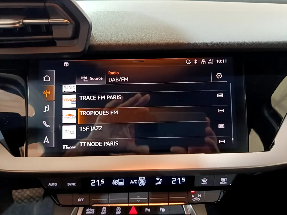 Écran tactile central de l'Audi A3 35 TFSI bleu, affichant les stations radio DAB/FM en intérieur.