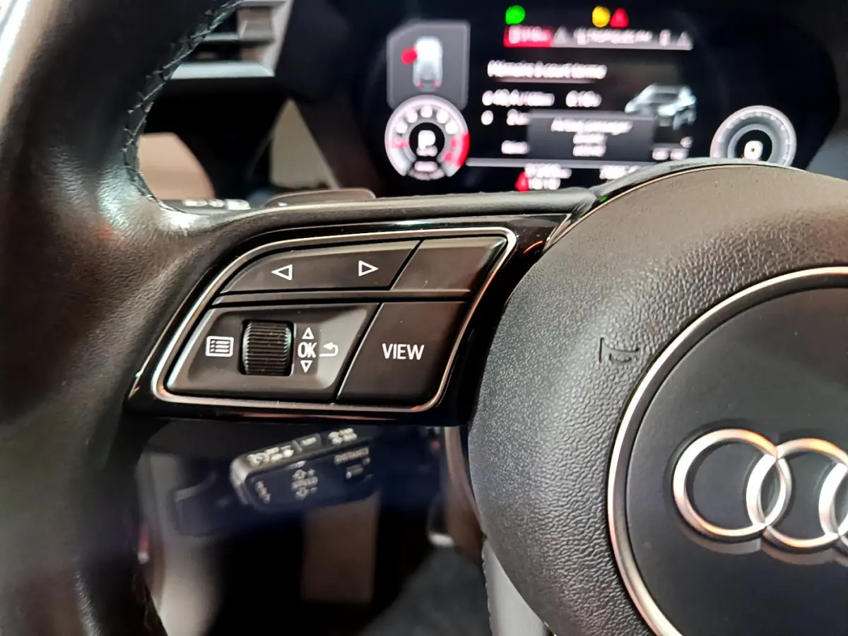 Gros plan sur la partie gauche du volant cuir d'une Audi A3 35 TFSI 2023 avec commandes multifonctions et tableau de bord numérique flou.