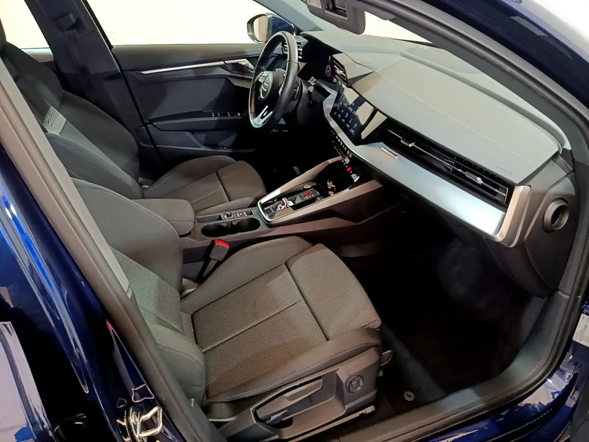 Intérieur de l'Audi A3 bleu 2023 vu côté conducteur, sièges tissu gris et tableau de bord moderne avec écran tactile.