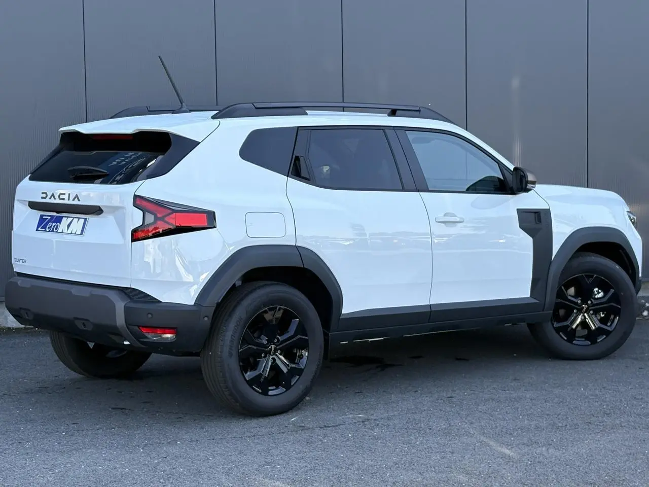 Vue 3/4 arrière droite du Dacia Duster 2026 blanc Glacier avec jantes noires et barres de toit noires.