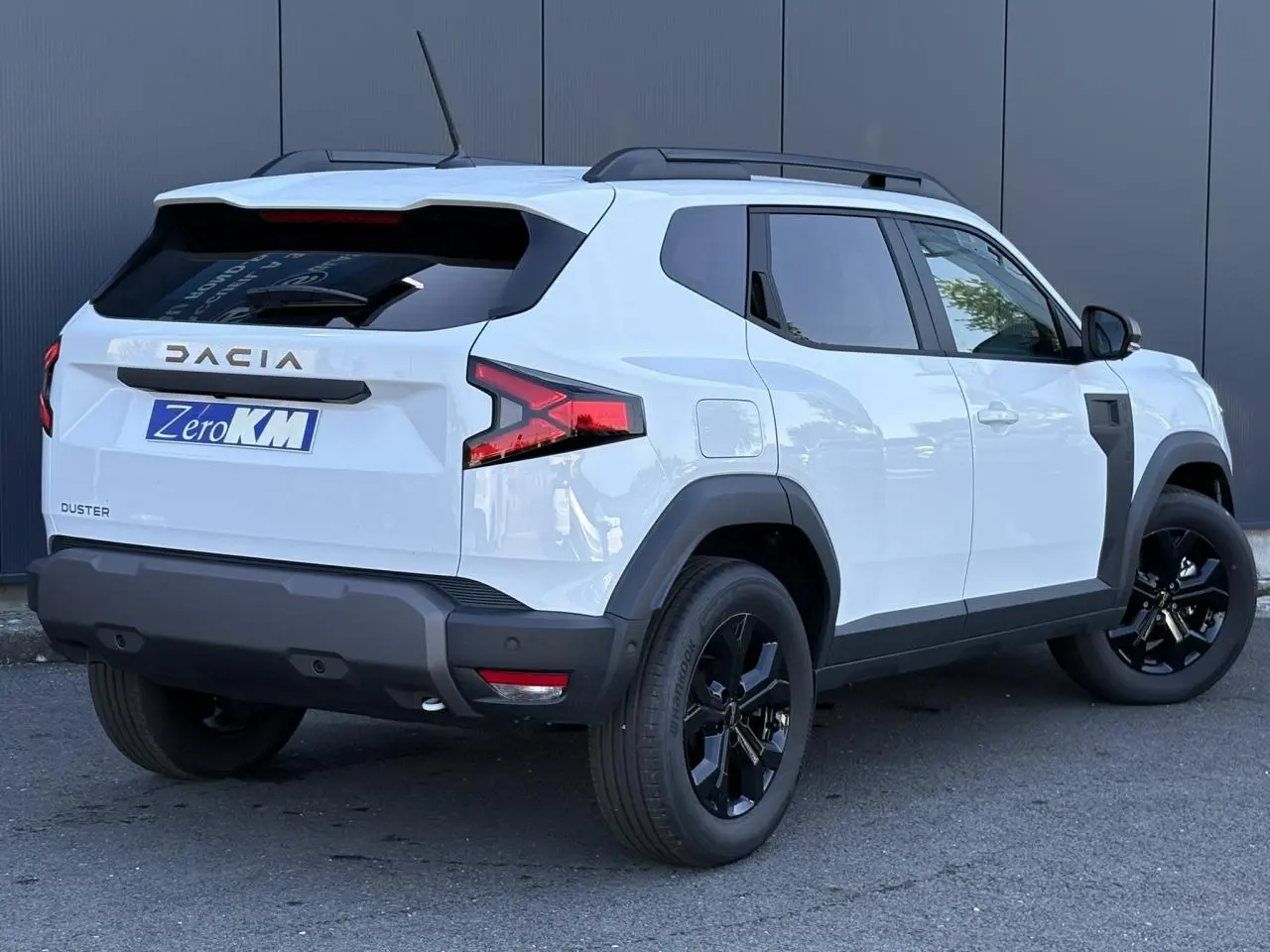 Vue 3/4 arrière droite du Dacia Duster 2026 blanc Glacier avec jantes alliage noires et barres de toit noires.