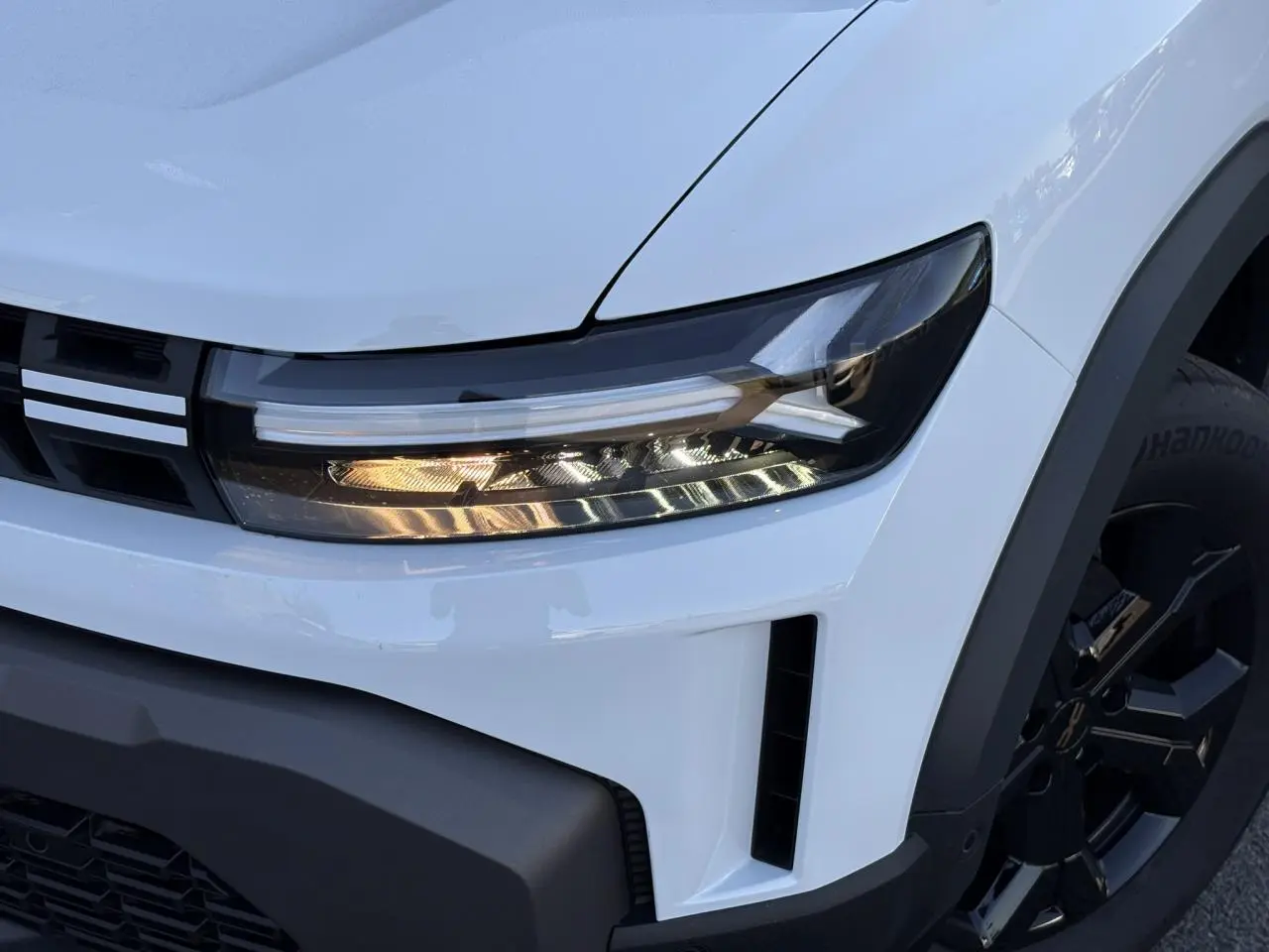 Gros plan sur le phare avant droit et la calandre du Dacia Duster blanc Glacier avec jante alliage noire.