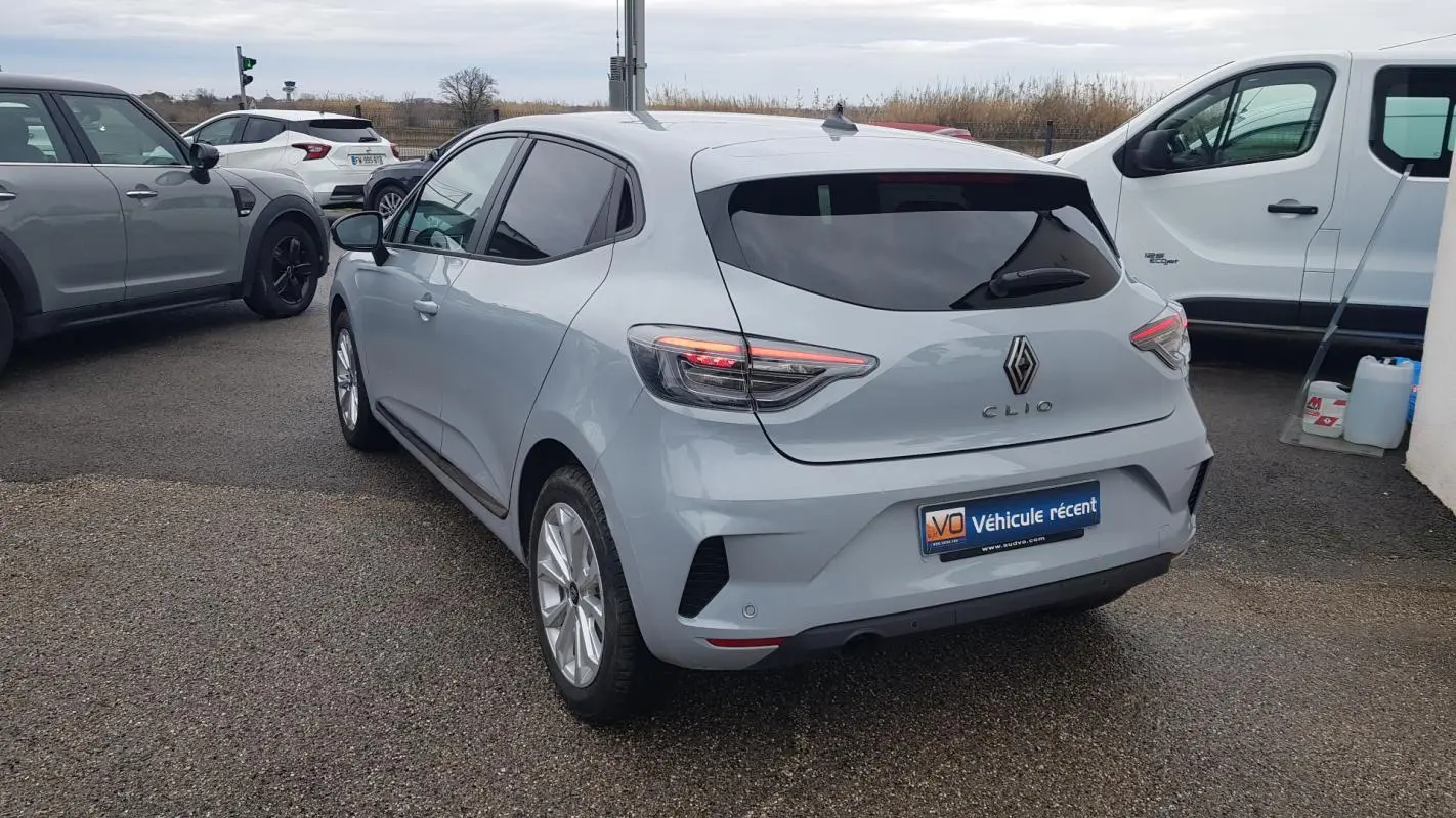 Vue 3/4 arrière droite d'une Renault Clio gris clair avec vitres surteintées et feux arrière allumés.