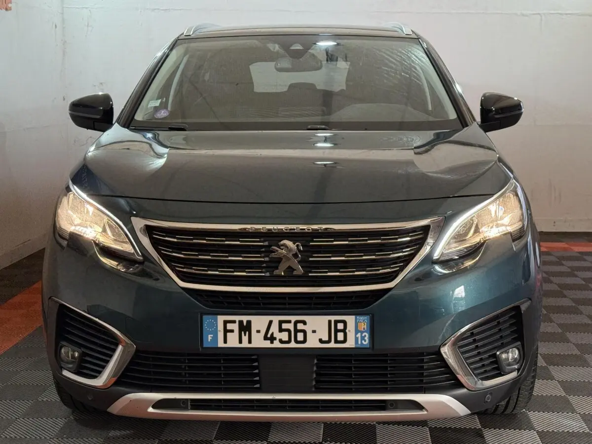 Vue avant d’un Peugeot 5008 bleu 2019 avec calandre chromée et phares allumés dans un garage.