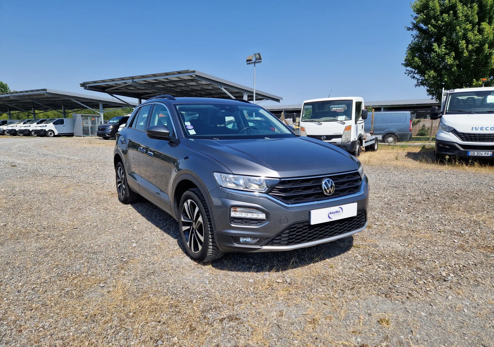 Volkswagen T-Roc gris foncé métal en 3/4 avant droit sur parking extérieur avec plusieurs véhicules utilitaires en arrière-plan.