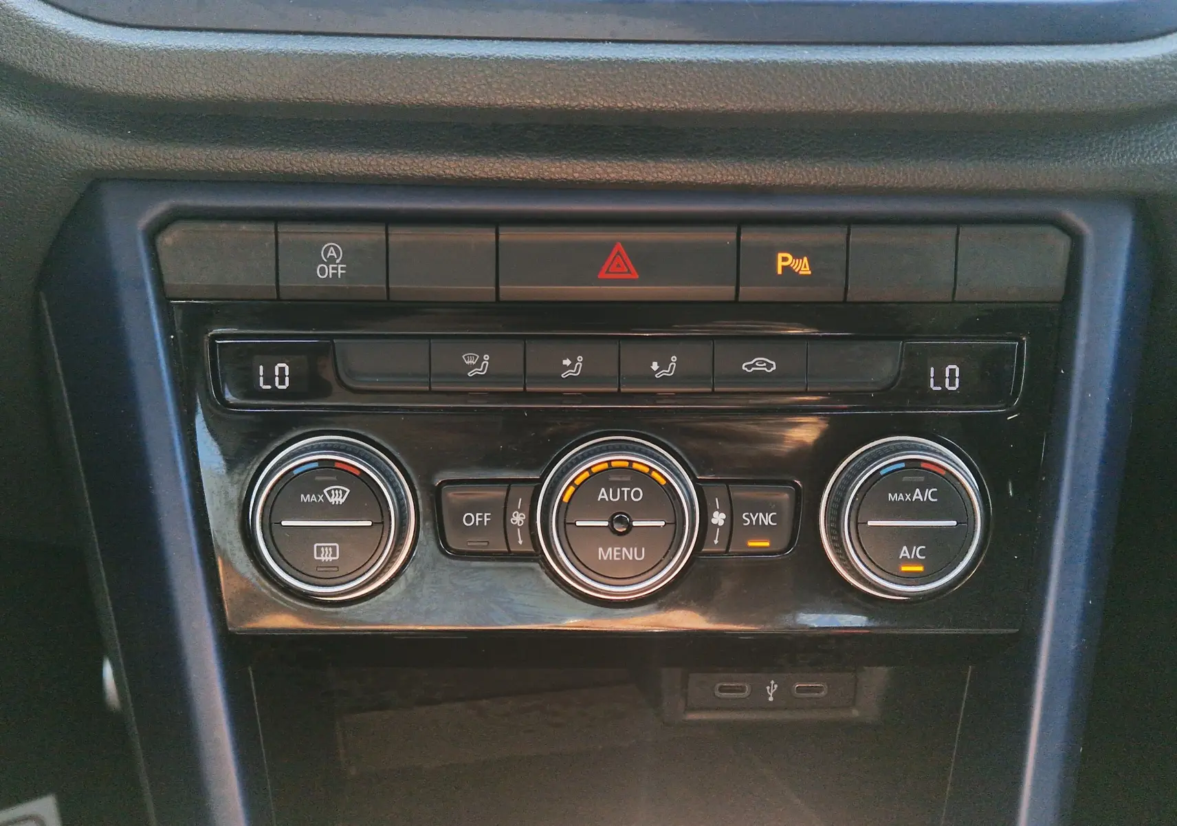 Gros plan sur la console centrale du Volkswagen T-Roc gris foncé, montrant les commandes de climatisation automatique et les boutons d’alerte.