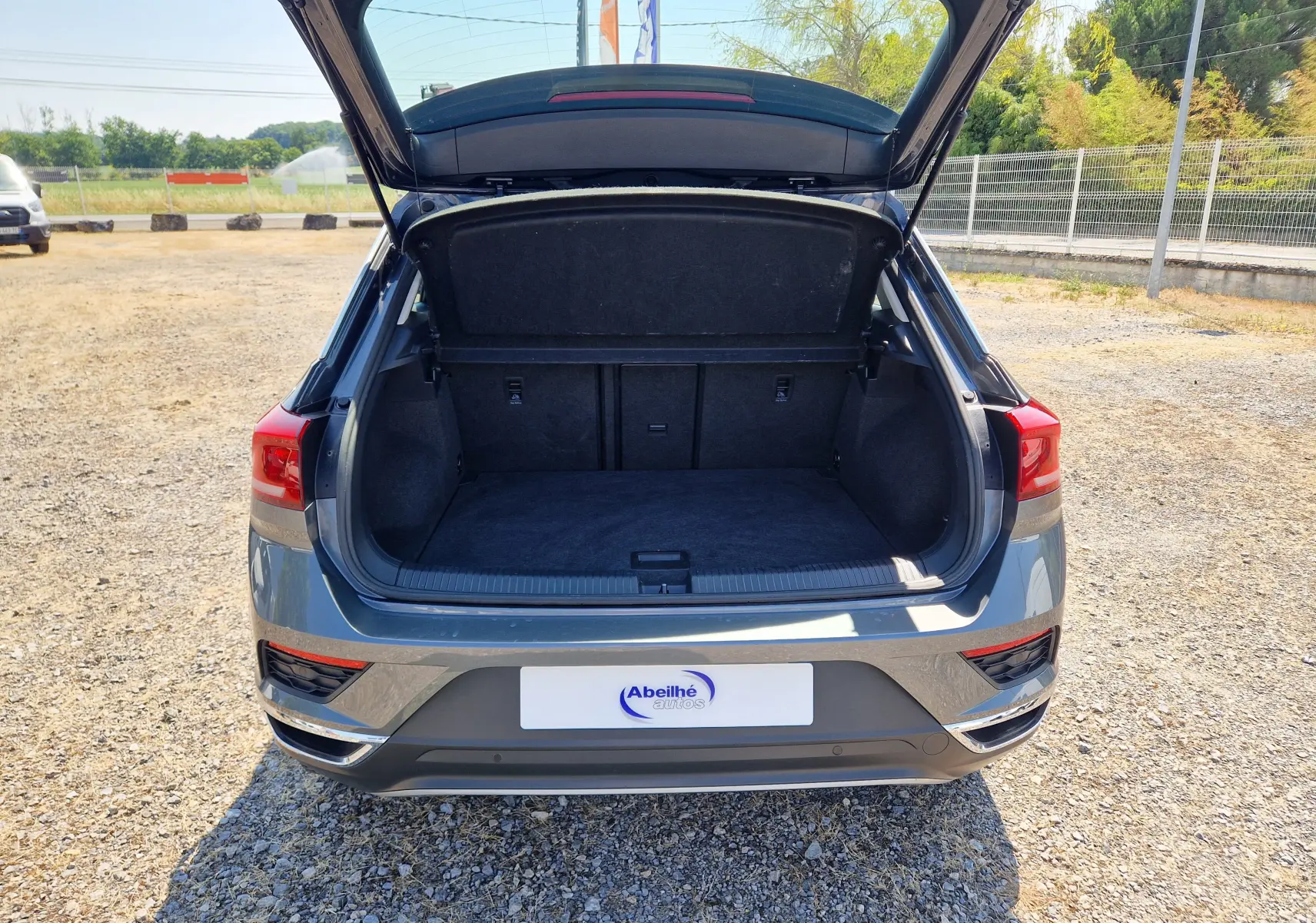 Vue arrière du Volkswagen T-Roc gris foncé métal 2021 avec coffre ouvert sur un sol gravillonné en extérieur.