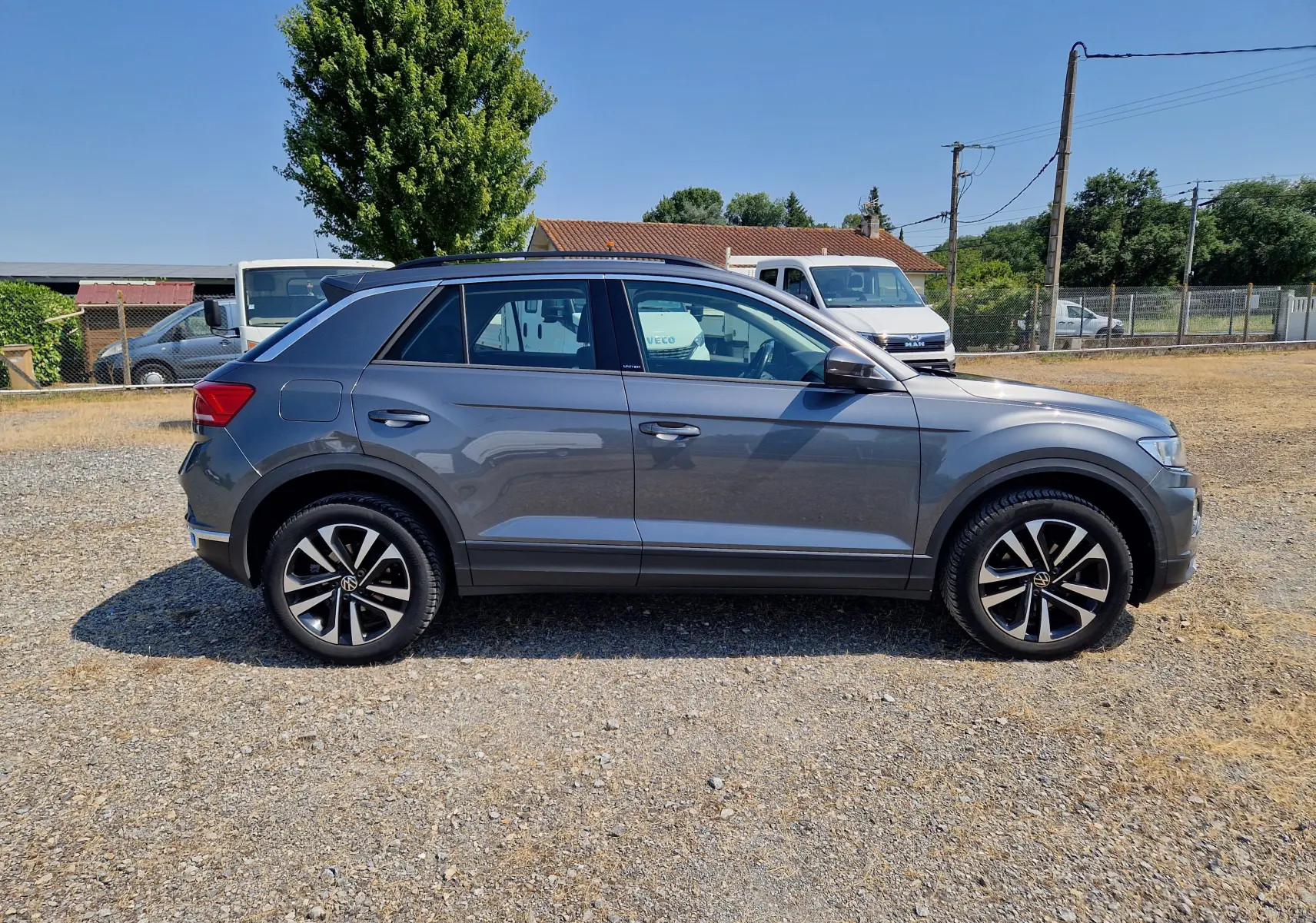 Profil côté droit du Volkswagen T-Roc 1.0 TSI 115 UNITED gris foncé métal, roues alliage 17 pouces, sur sol gravelé.