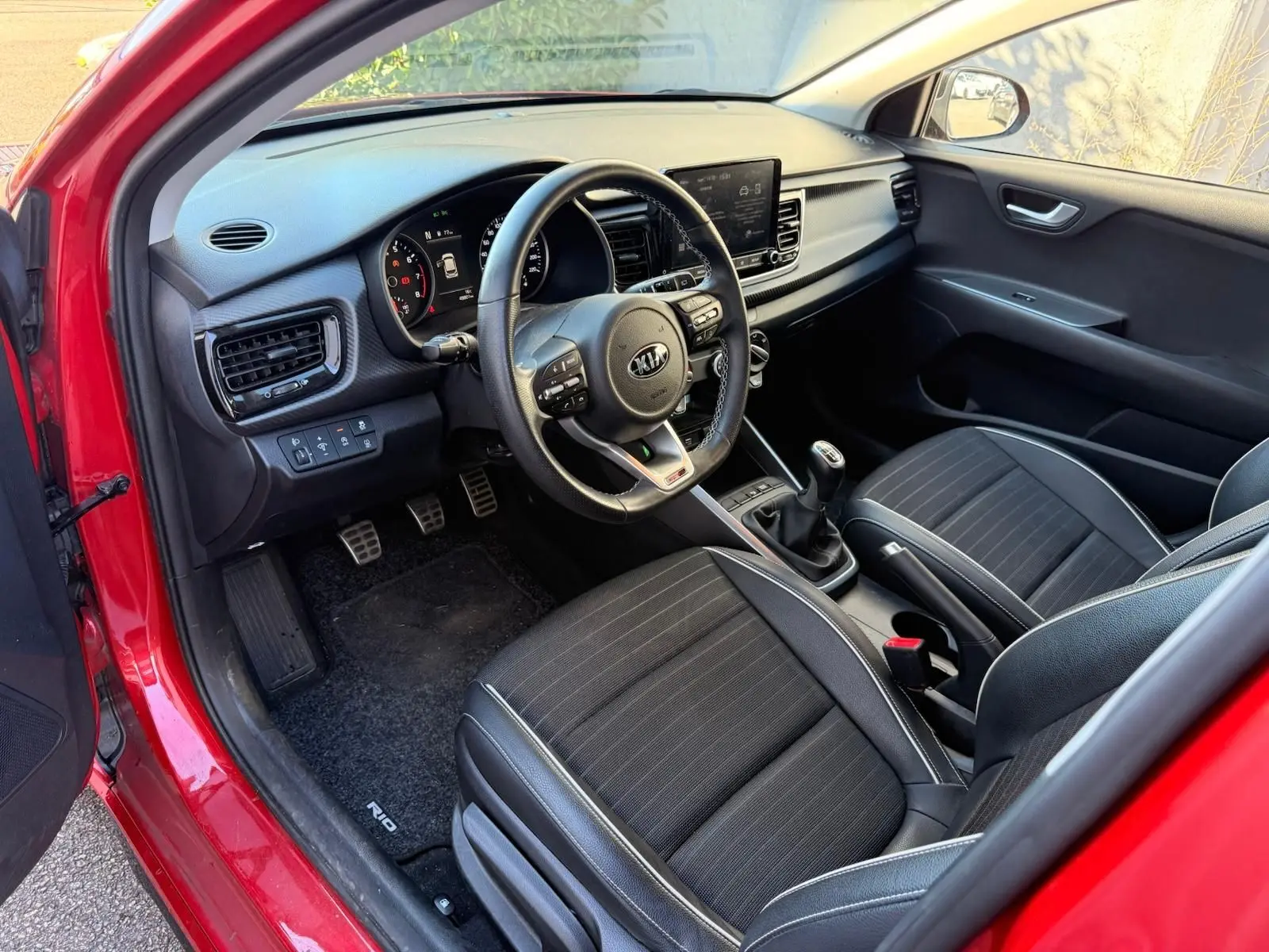 Intérieur côté conducteur d'une KIA RIO rouge grenadine 2022, montrant tableau de bord, volant multifonction et sièges noirs.